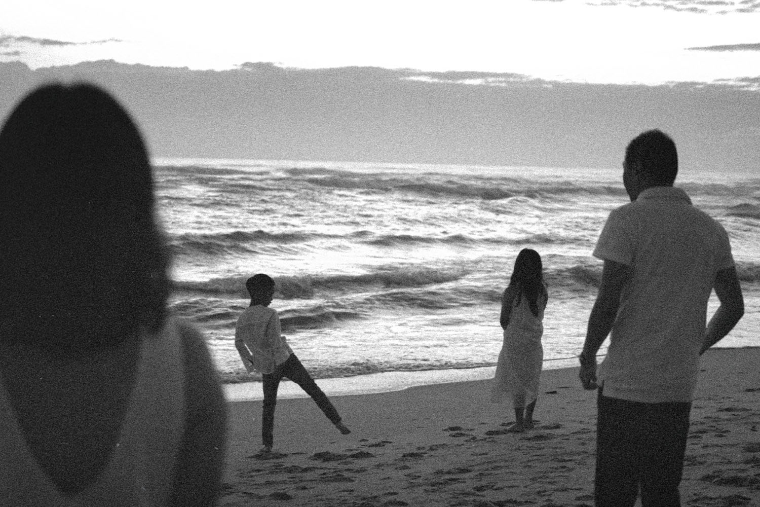A black-and-white scene of a family standing on a beach at dusk, watching waves roll onto the shore