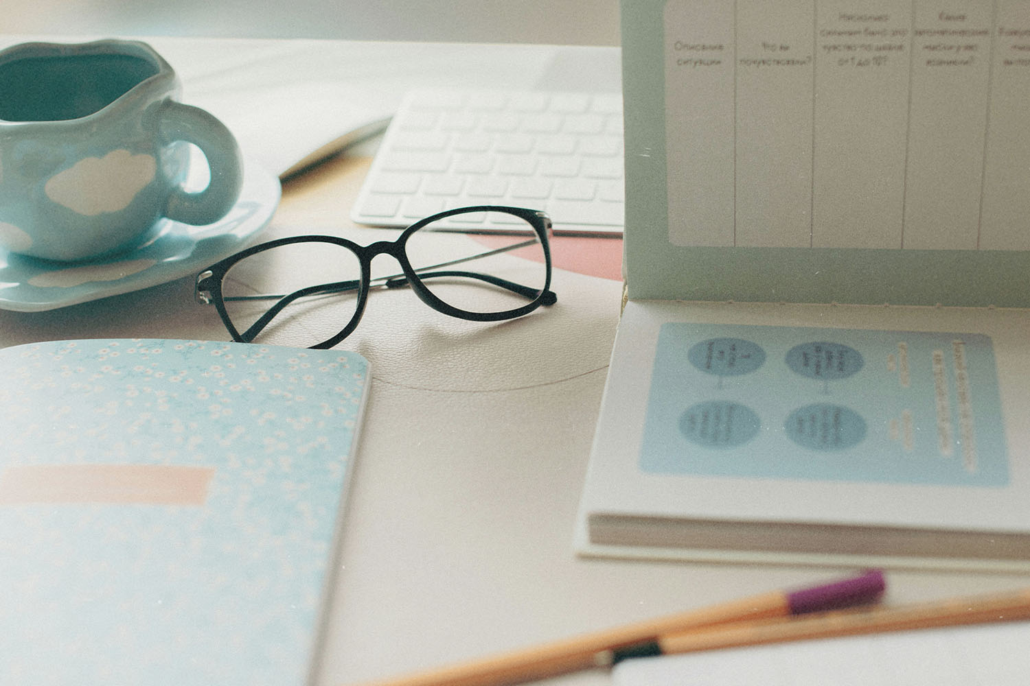 A workspace with glasses, notebooks, a planner, a keyboard, and a ceramic mug arranged neatly on a light-colored desk