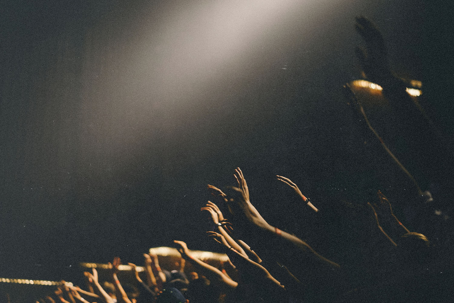 Numerous raised hands reach upward toward a bright spotlight in a dark room