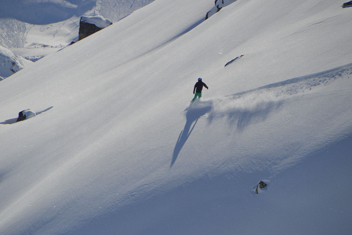 A skier glides down a wide, snow-covered mountain slope, leaving a trail of powder behind under bright sunlight