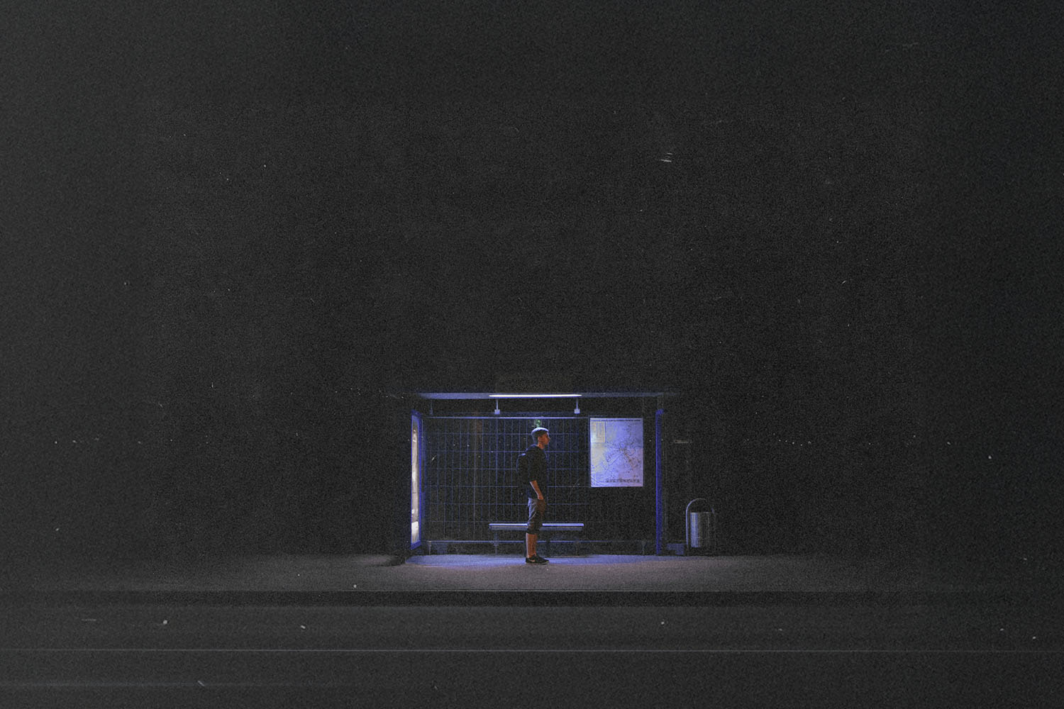 Single person stands under the dim blue light of a bus stop shelter at night, surrounded by darkness