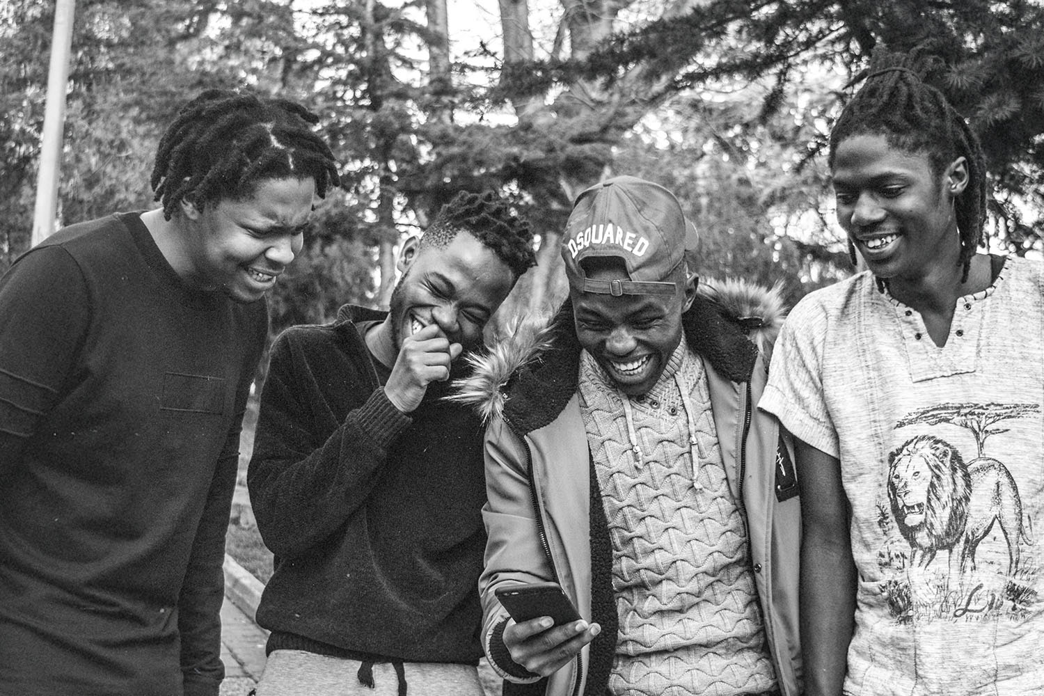 Four people stand outdoors closely together, looking at a smartphone one person is holding