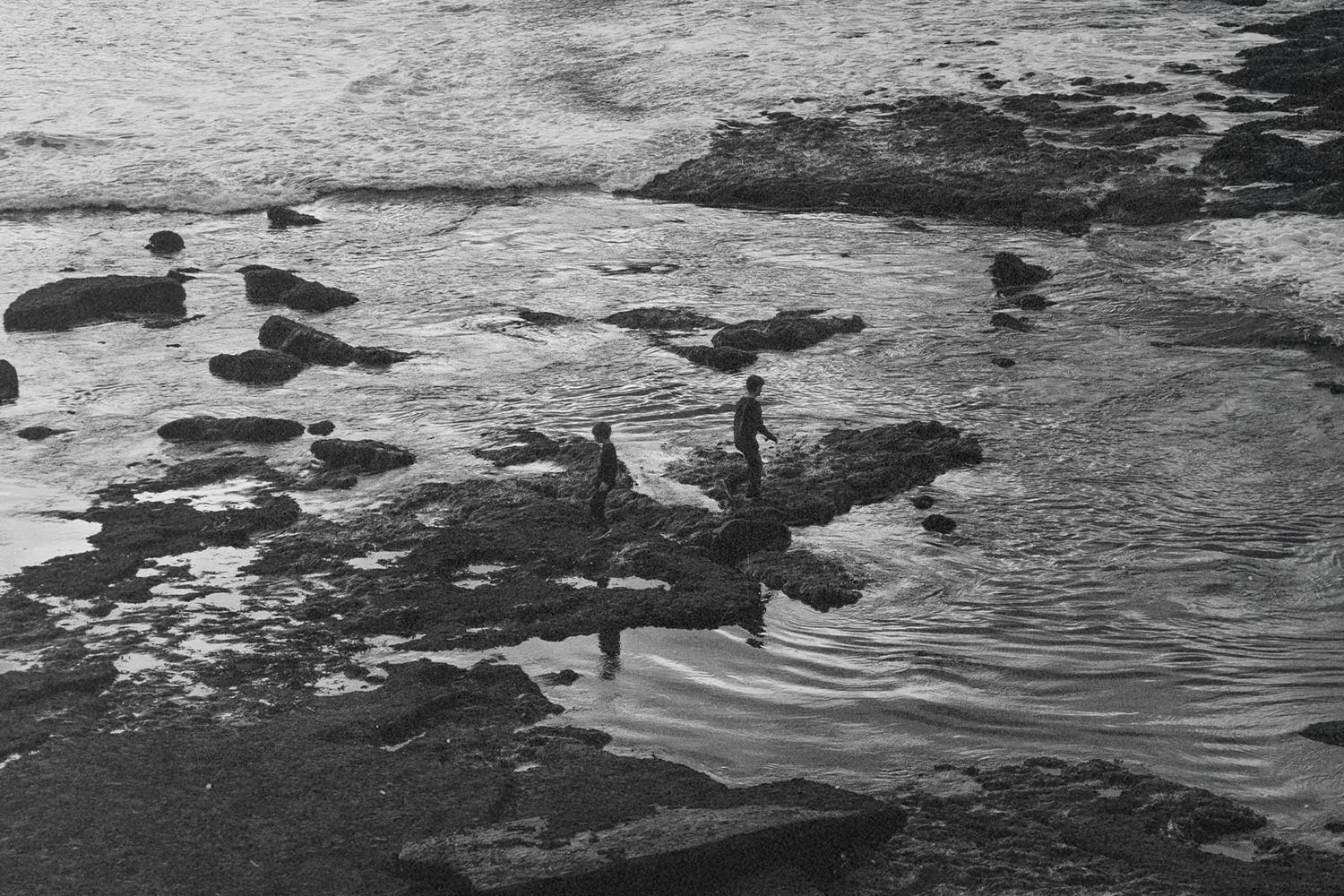 Two children walk along rocky tide pools at the edge of the ocean as waves roll in