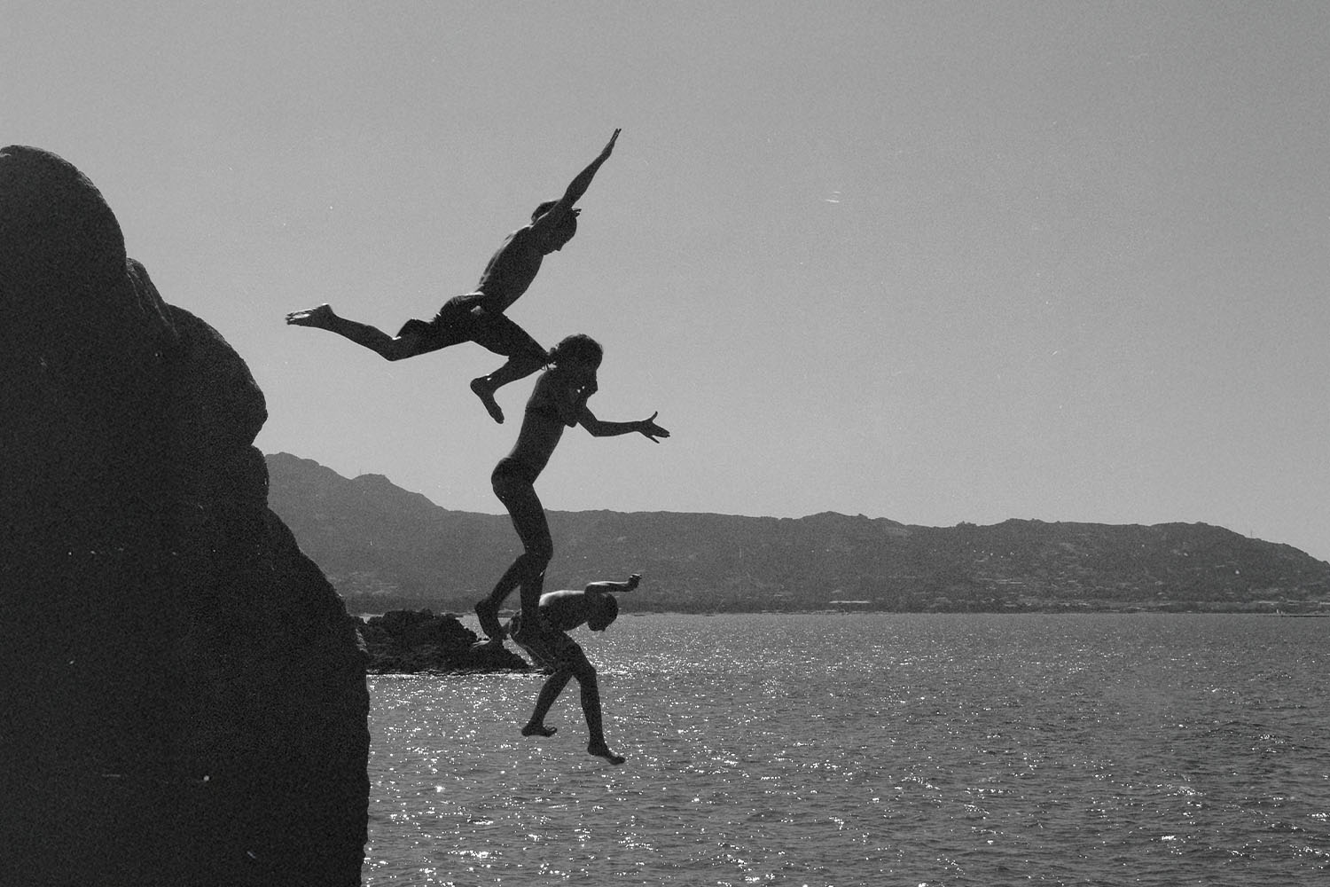 Three people leap from a rocky cliff into the water below, captured mid‑air in a black‑and‑white scene