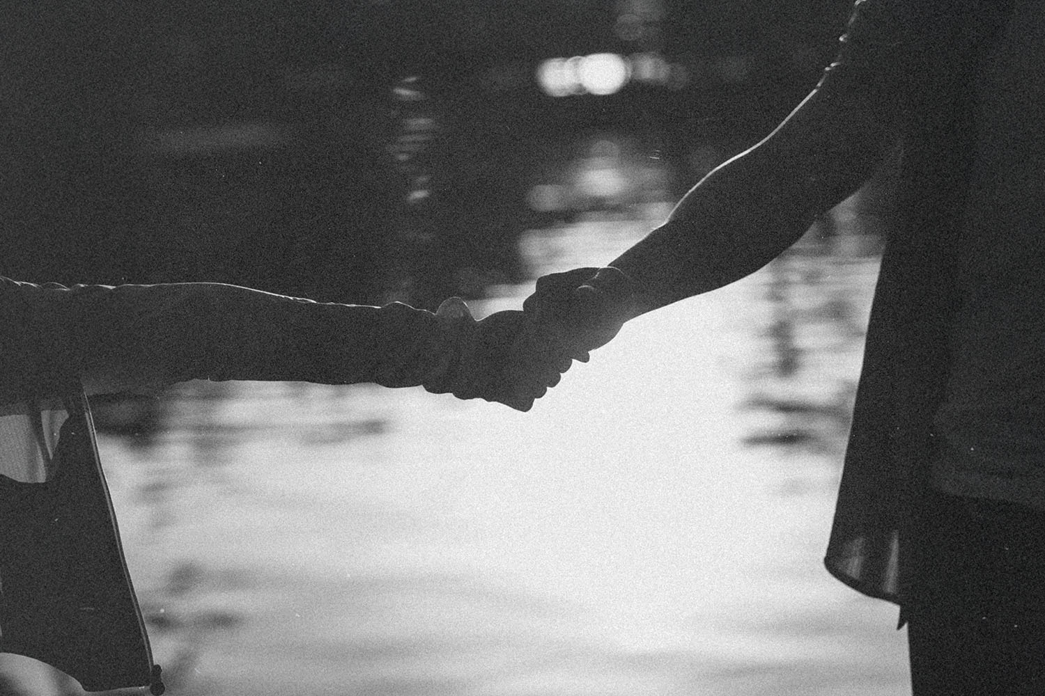 Two people hold hands beside a reflective body of water, shown as a soft black‑and‑white silhouette