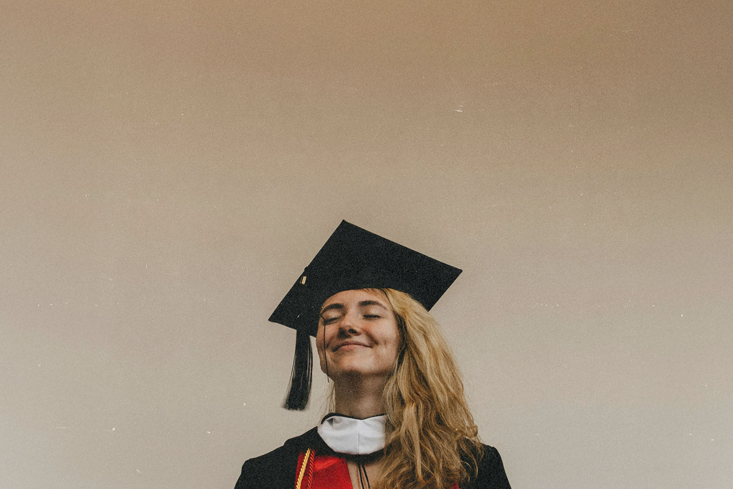 A graduate wearing a cap and gown stands against a plain beige background