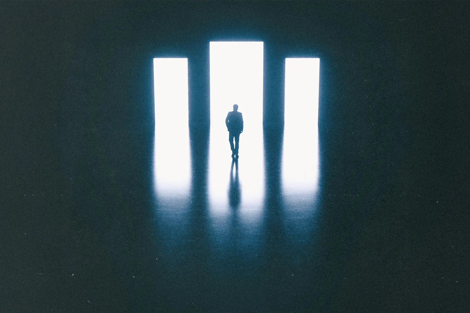 Silhouette of a person standing before three tall, bright rectangular doorways in a dark room, with light reflecting across the floor