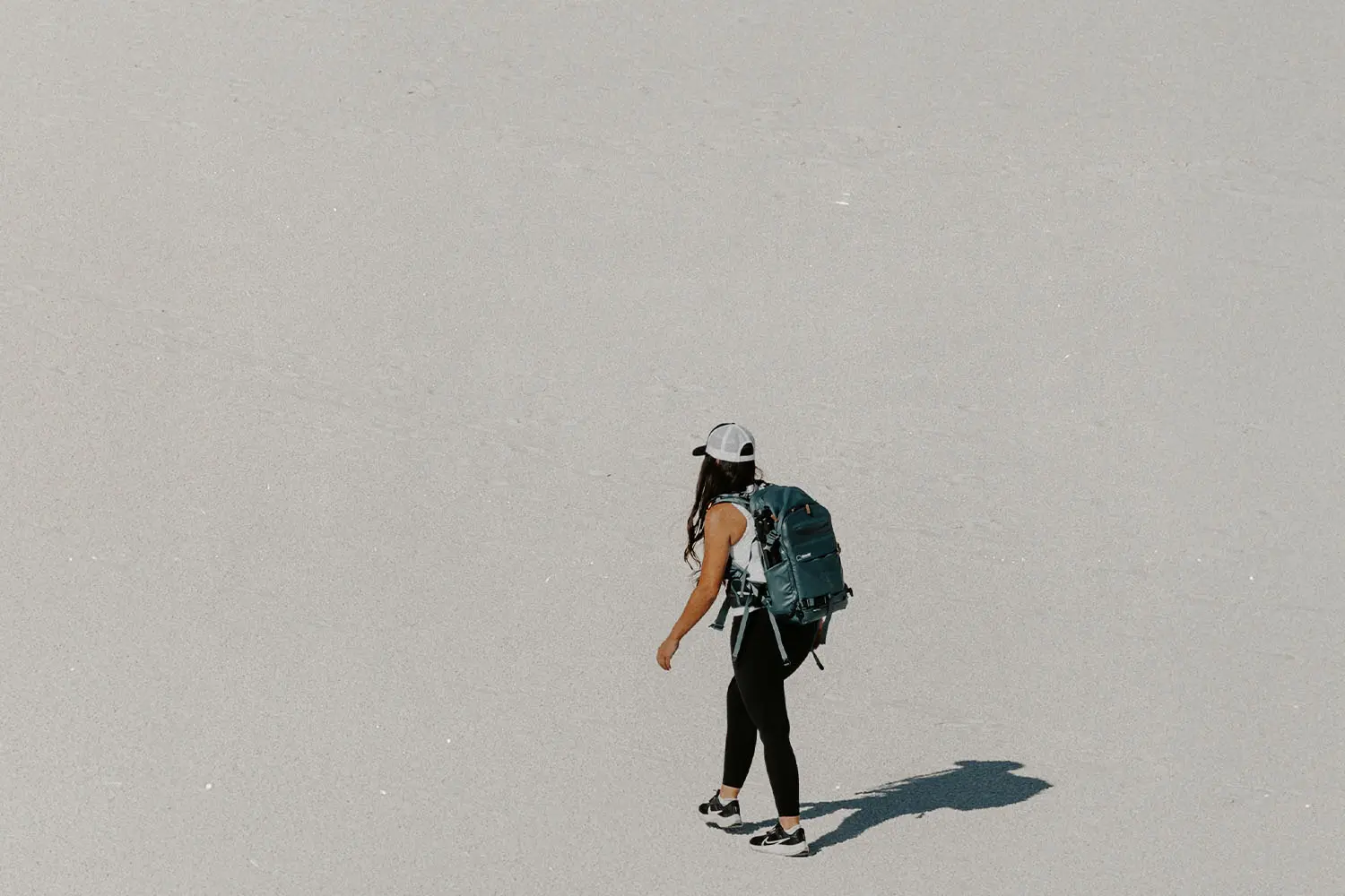 Person wearing a backpack walking alone across a wide, bright, sandy landscape, casting a long shadow in the sun