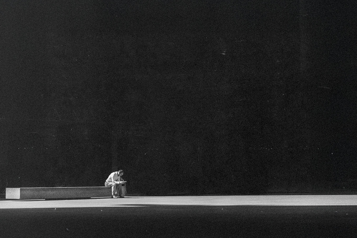 Person sitting alone on a long bench in a vast, dimly lit space, leaning forward with their head down