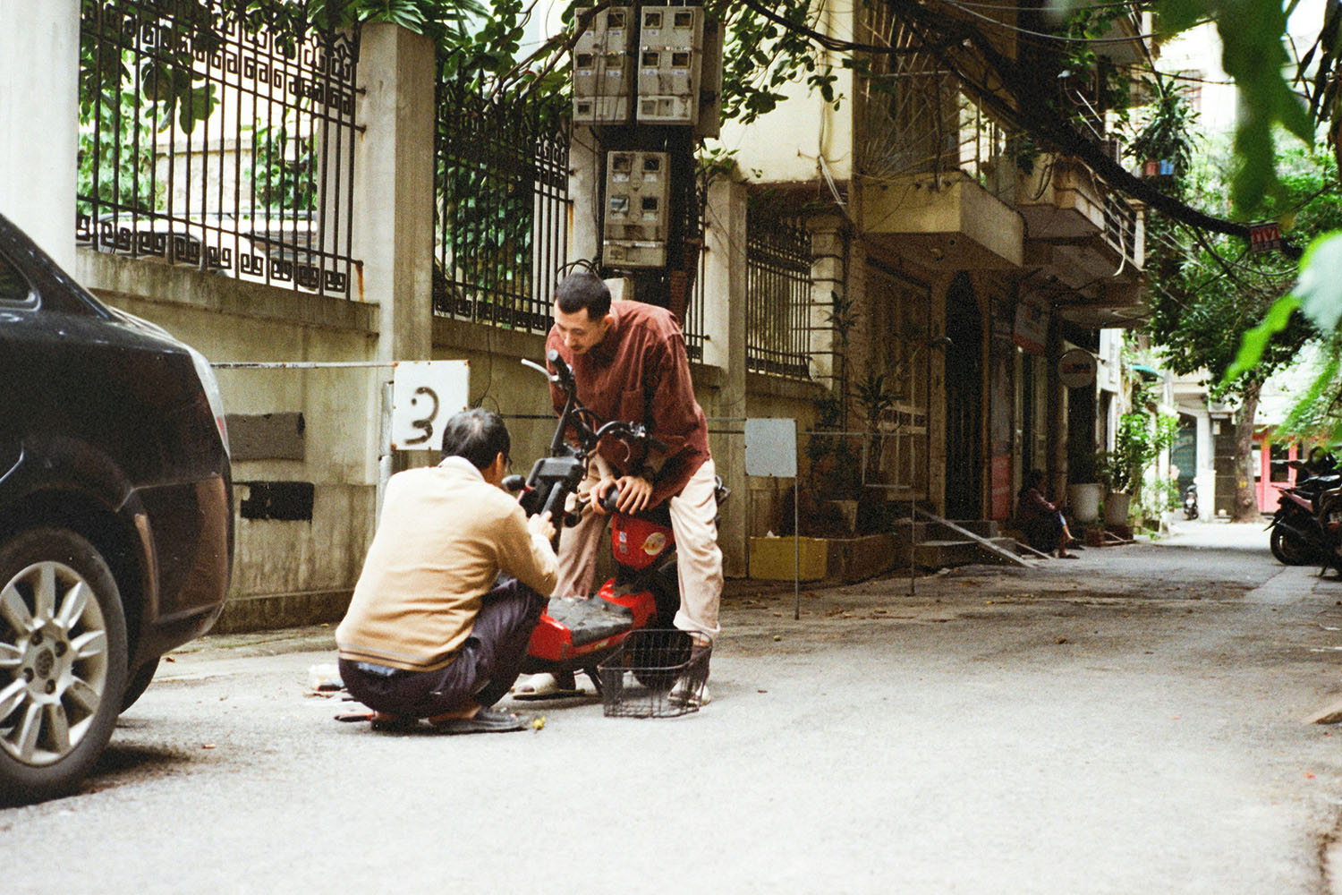 Two people working together to repair a red motorbike on a narrow residential street lined with cars, plants, and aging buildings