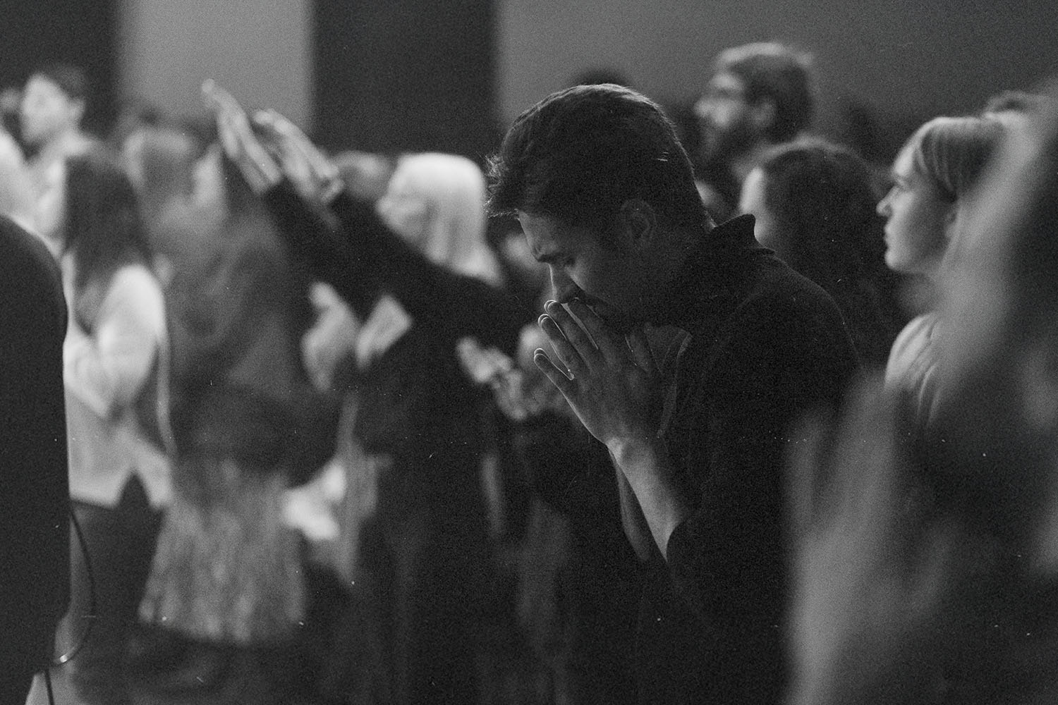 Group of people standing closely together in a dimly lit room with several individuals raising their hands and one person in the foreground holding their hands together in front of their face