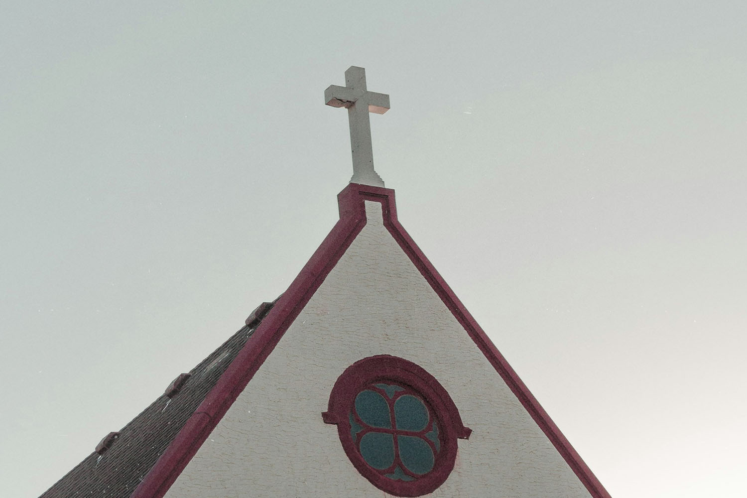 Steeple of a small church with a white cross on top and a circular stained‑glass window, set against a pale sky