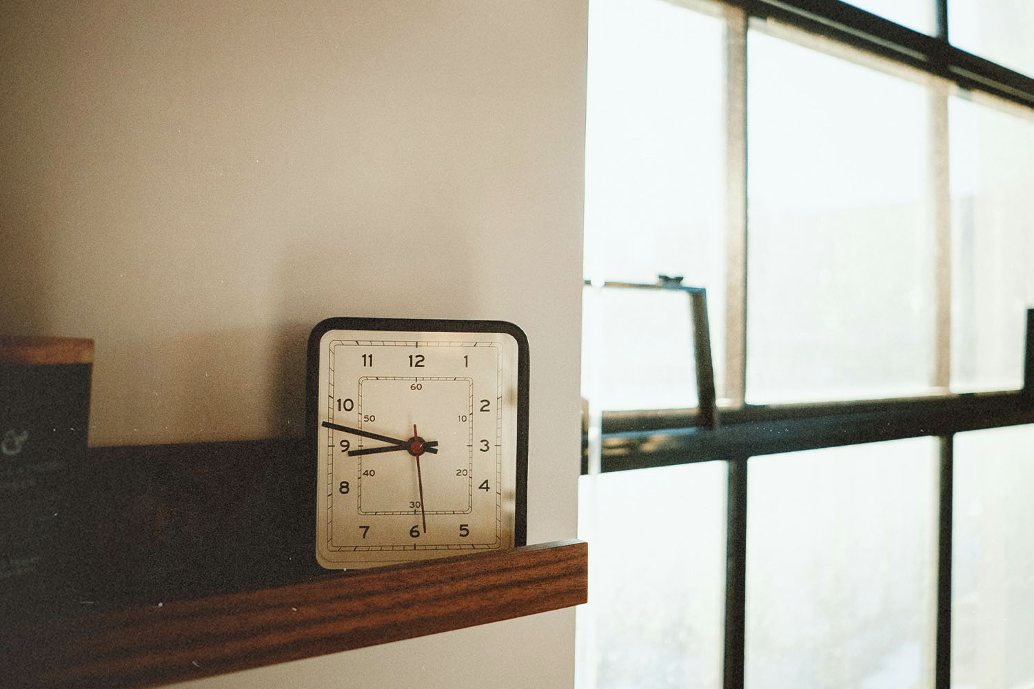 Square analog clock sitting on a wooden shelf next to a large sunlit window with metal frames