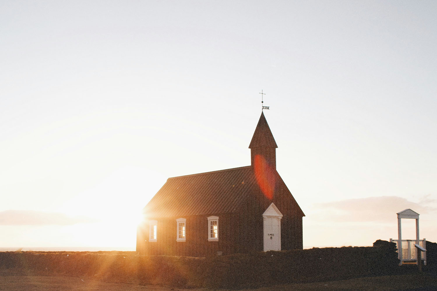Small dark wooden church standing in an open landscape at sunrise, with warm sunlight shining behind the building and a clear sky above