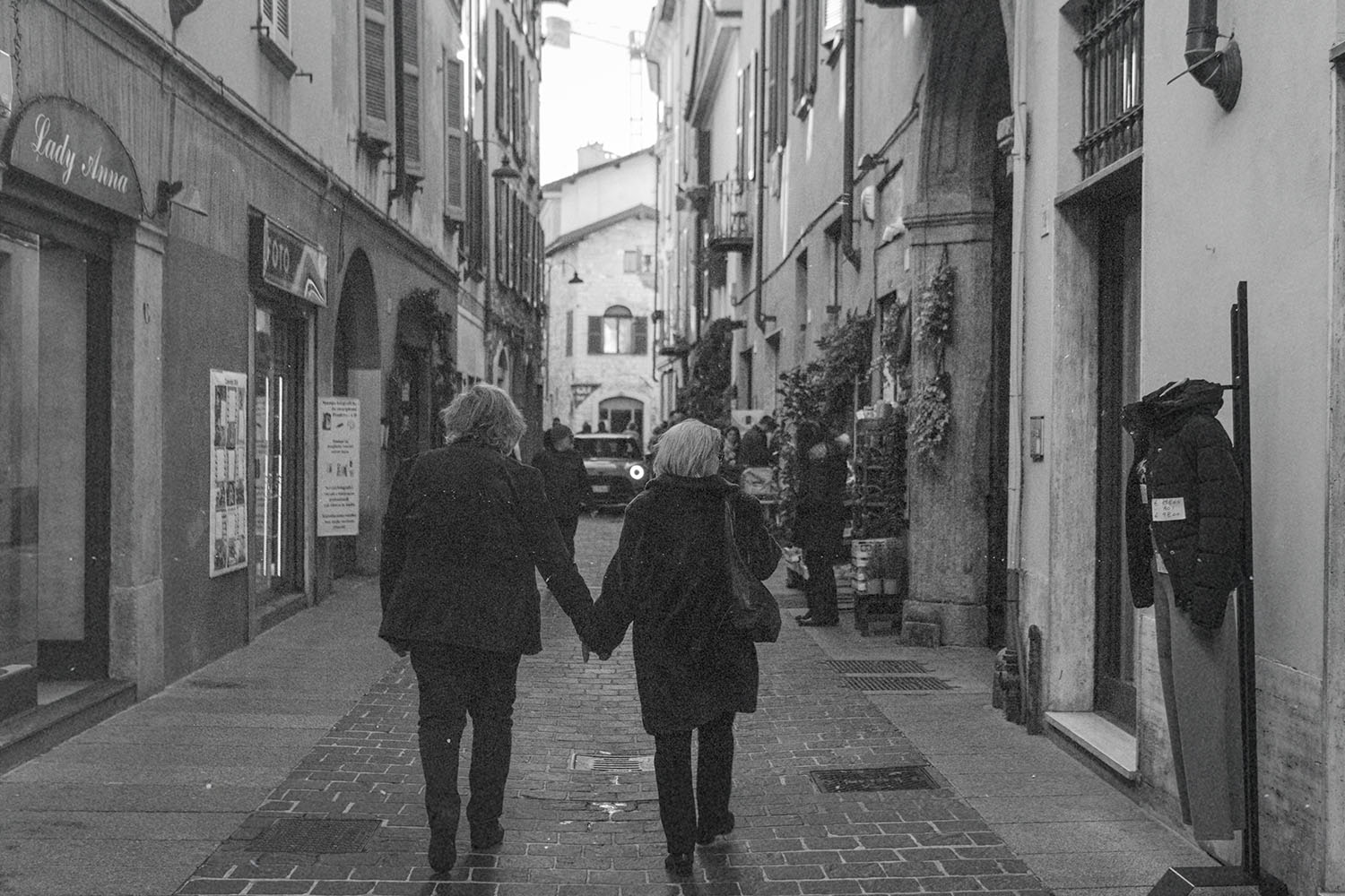 Two people walking hand‑in‑hand down a narrow cobblestone street lined with shops and tall historic buildings