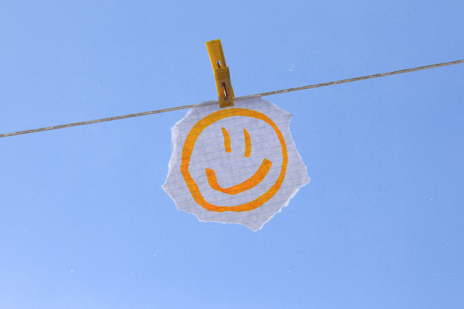 Hand‑drawn smiley face on a torn piece of paper clipped to a clothesline against a clear blue sky