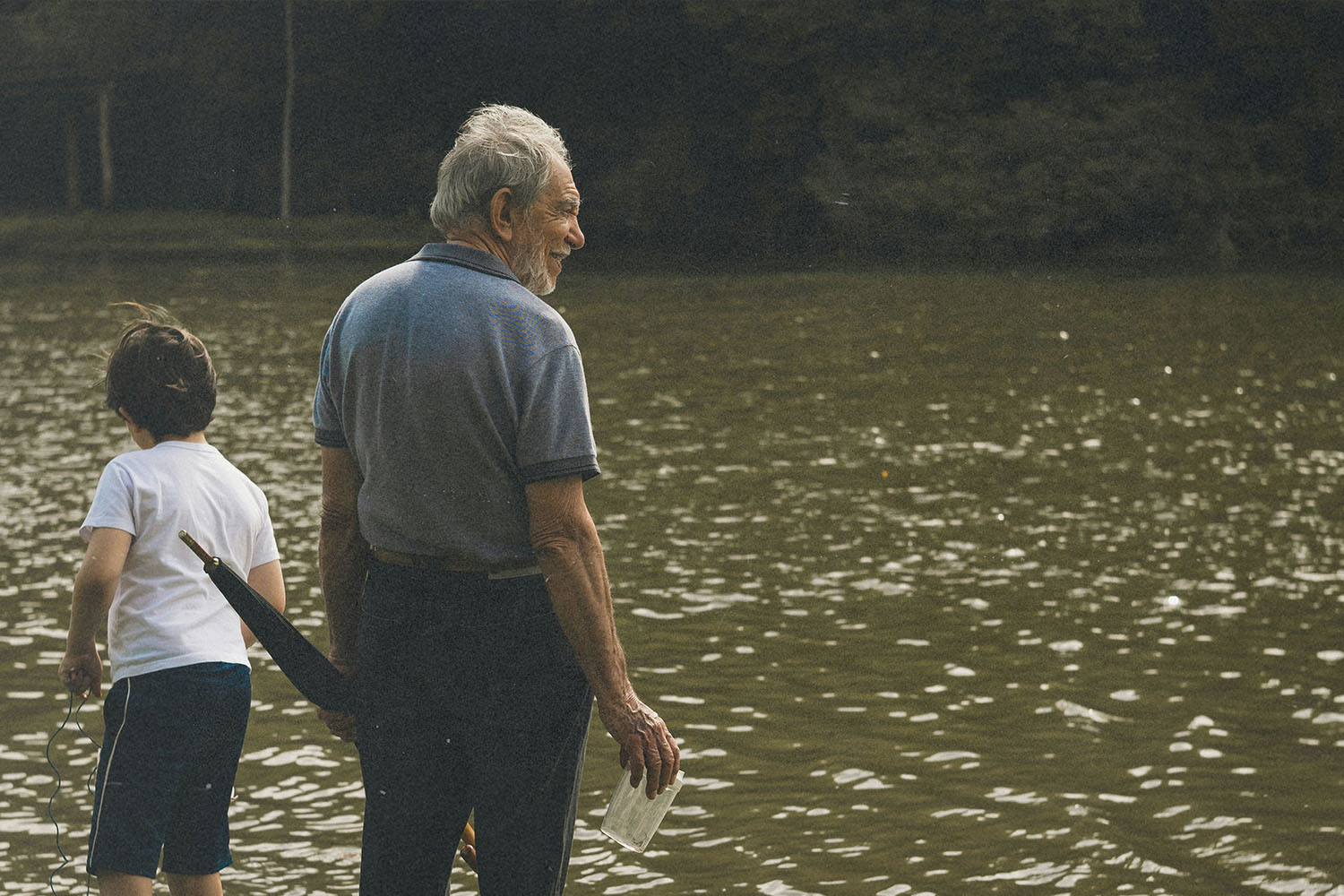 Older adult and child standing at the edge of a calm, reflective body of water, holding items in their hands while looking out across the surface