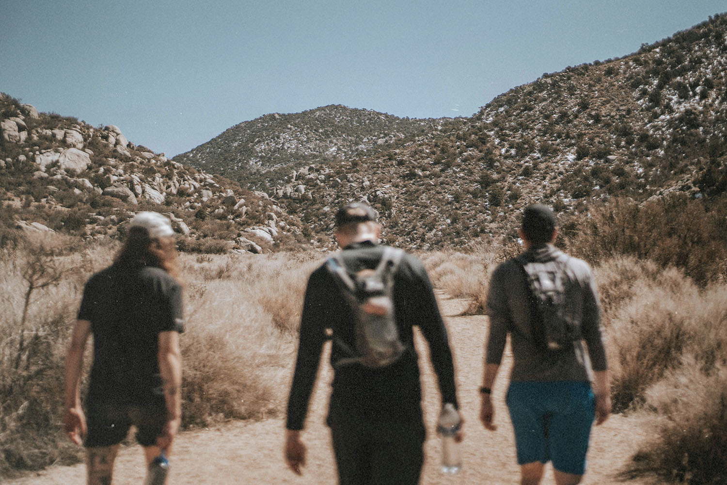 Three people walk along a dirt trail surrounded by dry brush, with rocky mountains rising in the background under a clear sky