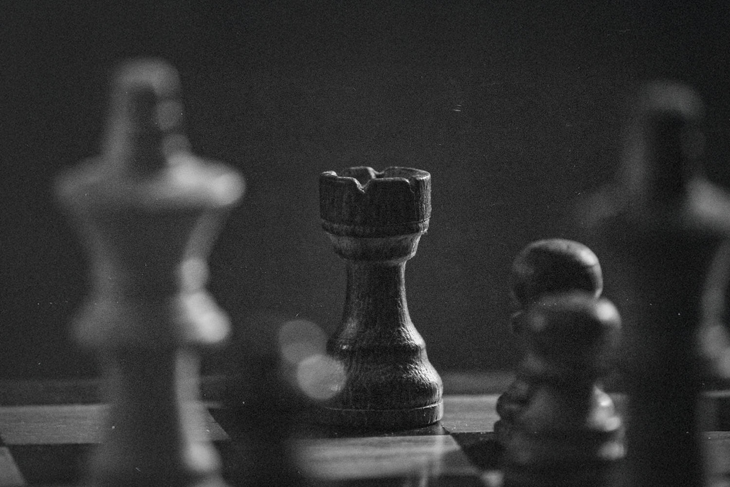 Black-and-white photograph of a chessboard with a wooden rook in sharp focus among blurred pieces
