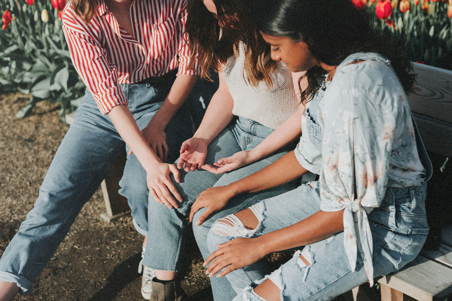 Three people sit together outdoors, hands lightly touching in a supportive gesture, surrounded by sunlight and nearby tulips