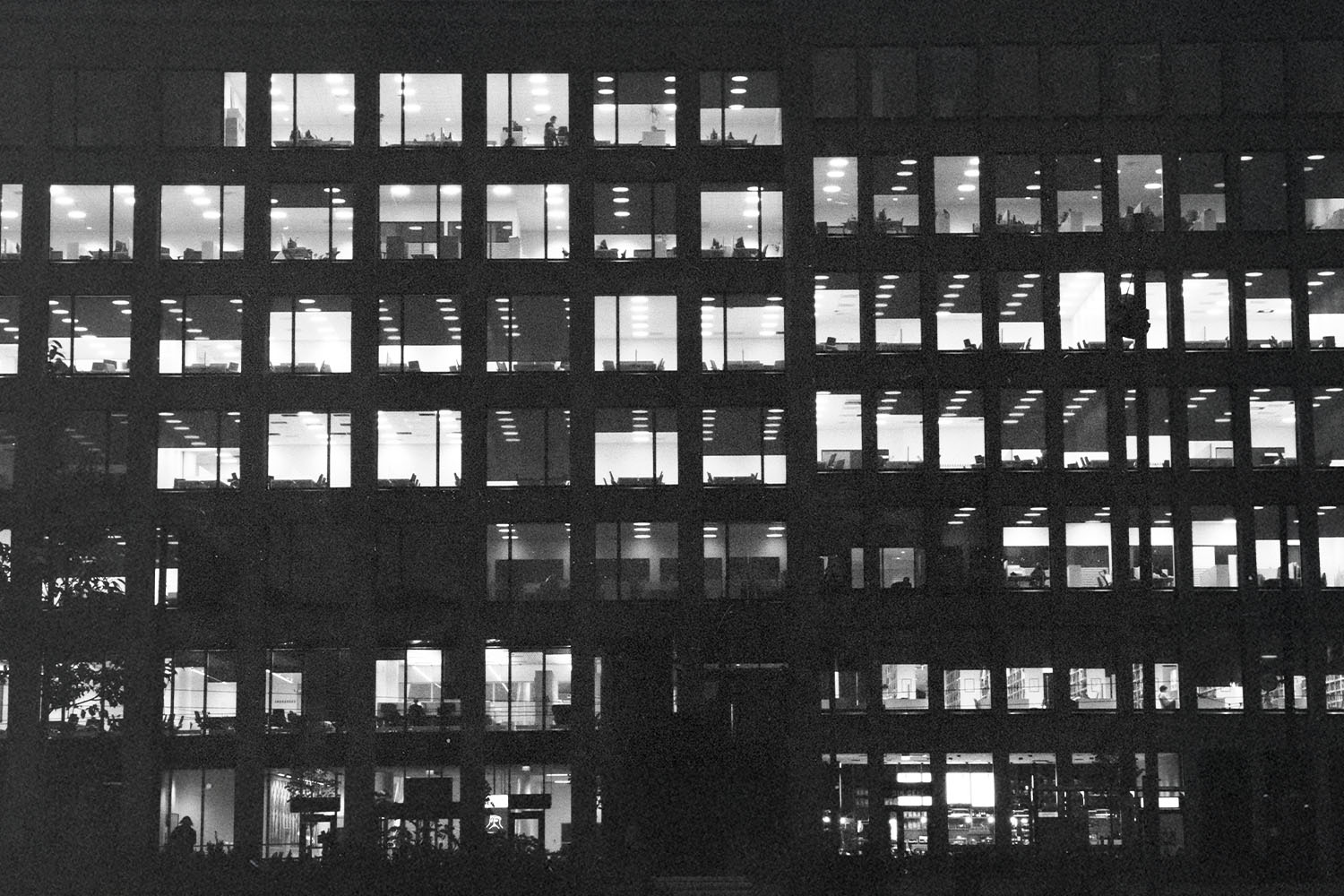 A nighttime view of a large office building, its grid of windows illuminated to reveal people working inside