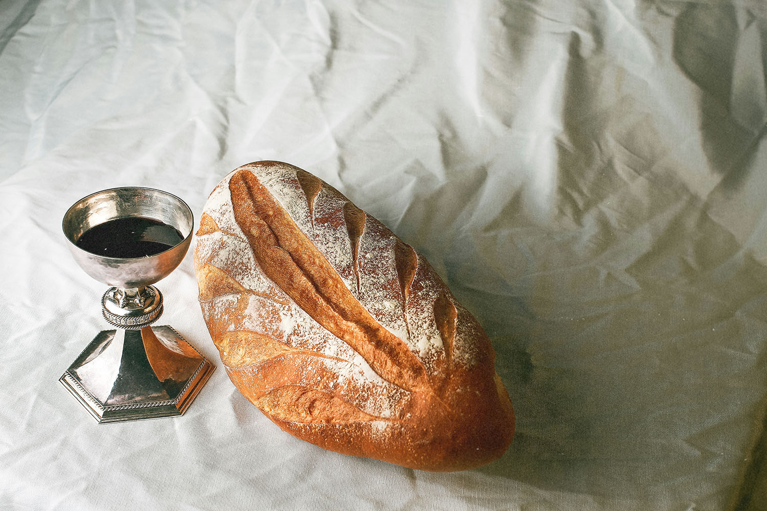 Loaf of rustic bread sits beside a silver communion cup filled with dark liquid on a softly wrinkled white cloth