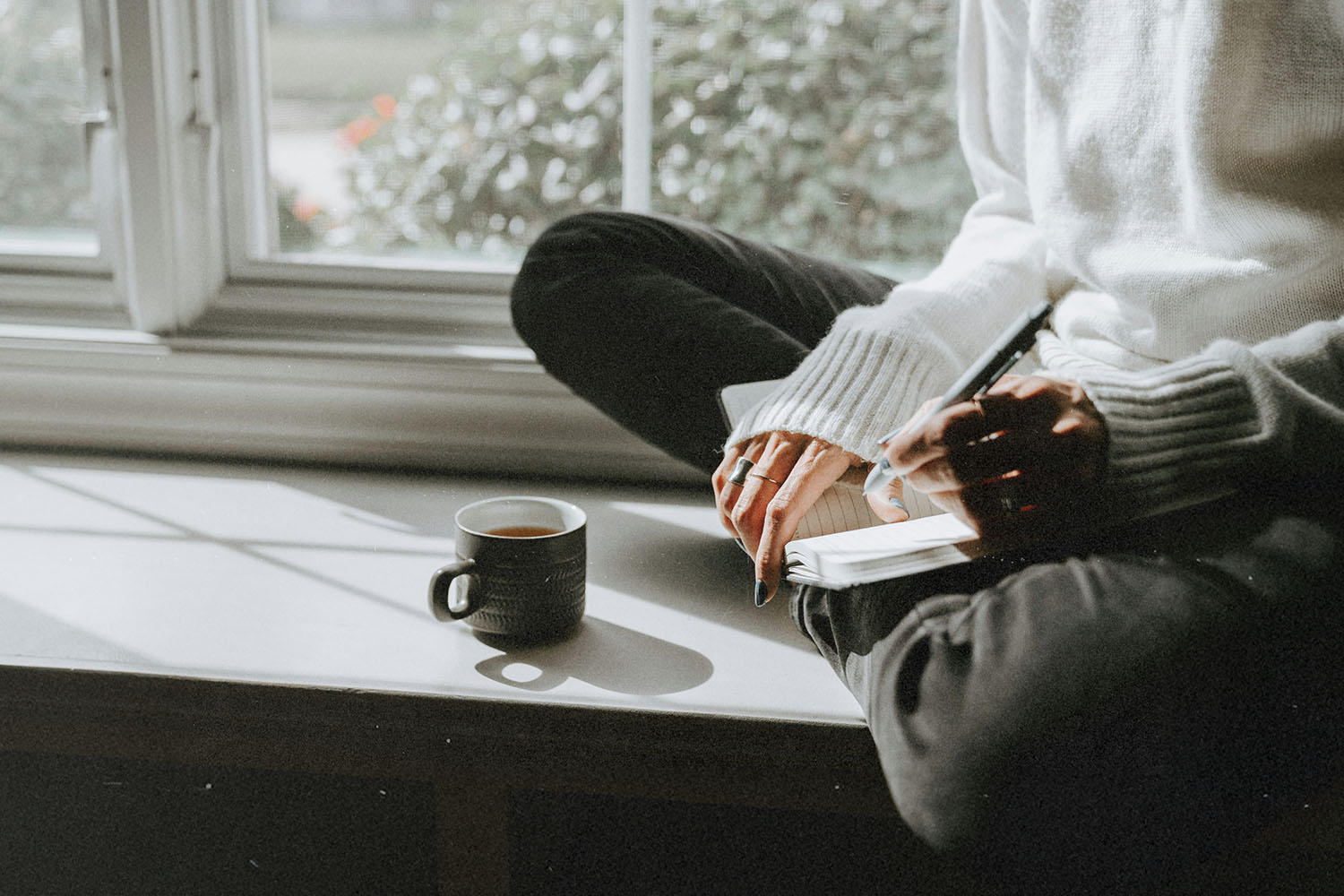 Person sits cross‑legged on a windowsill, writing in a notebook beside a mug of coffee in the sunlight