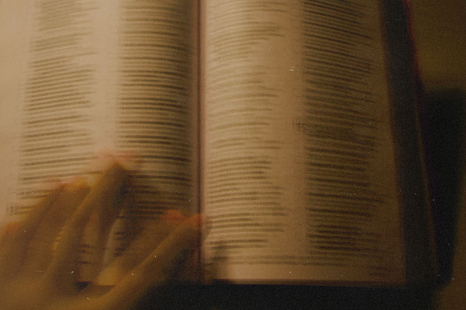 A blurred close‑up of an open Bible with a hand gliding across the page, softly lit in warm tones