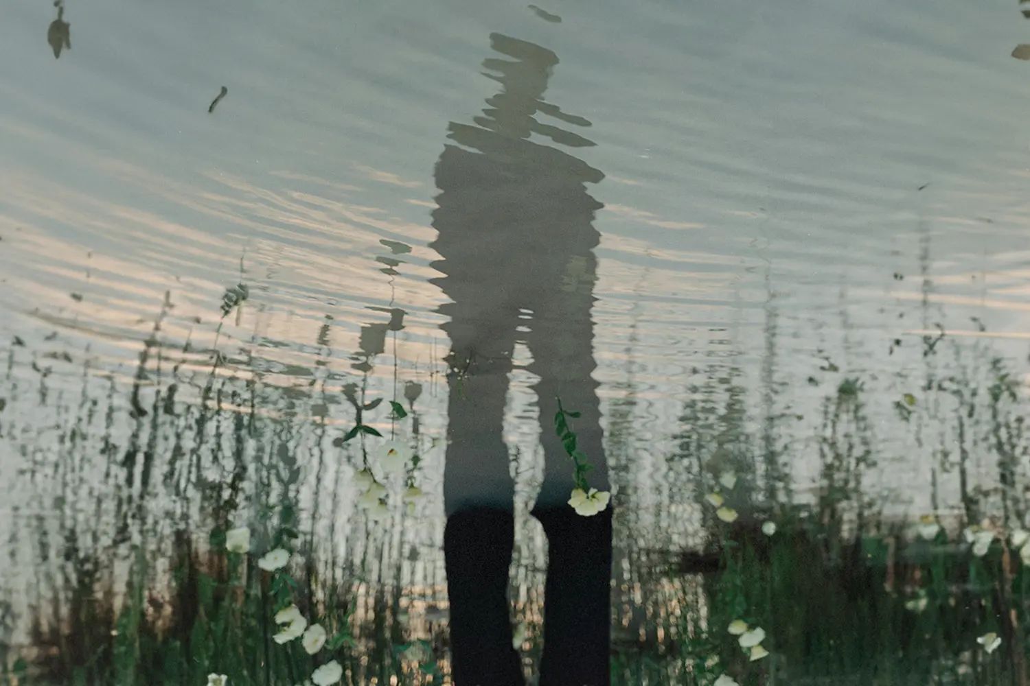 Reflection of a person standing beside rippling water with grass and white flowers, symbolizing Lent’s themes of reflection, prayer and spiritual renewal.