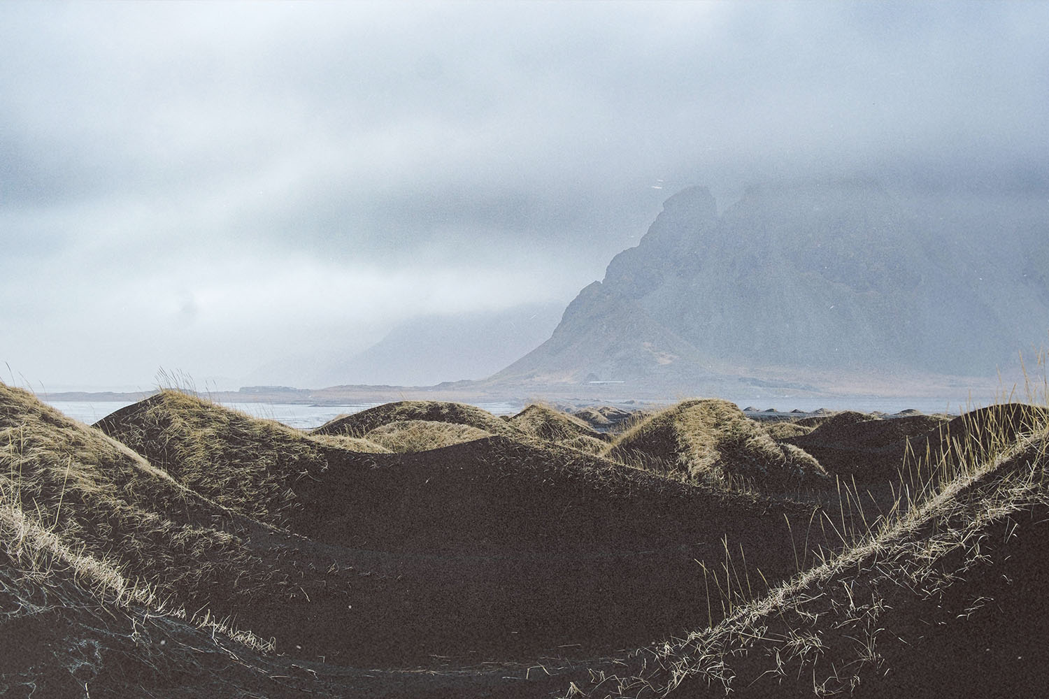 Wind-shaped black sand dunes under a cloudy sky with a mist-covered mountain in the distance
