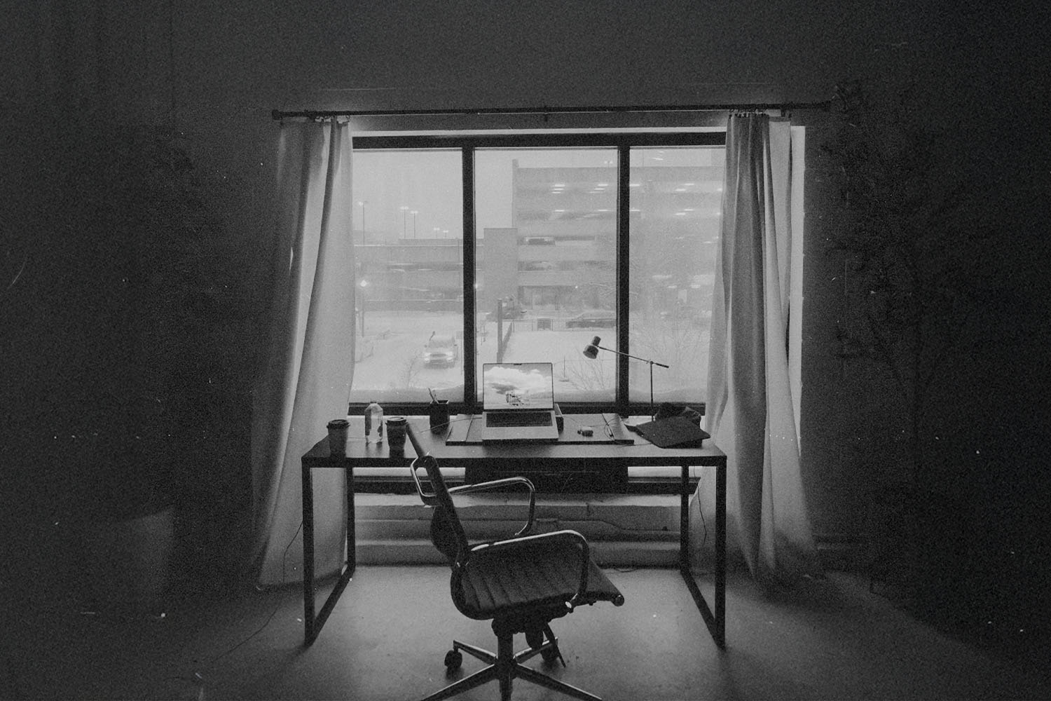 Simple office space with a laptop and lamp on a desk framed by open curtains and an urban view