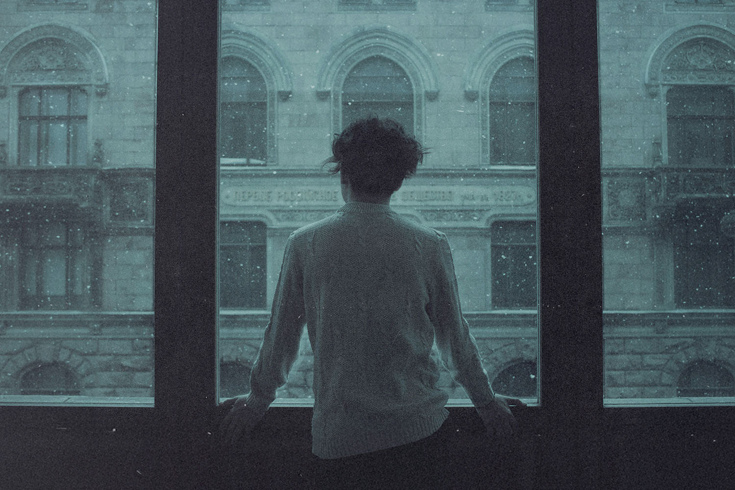 A person stands at a large window, looking out at an ornate stone building as snow falls outside