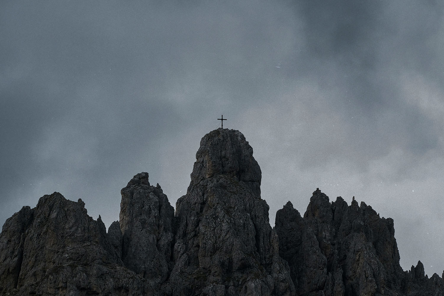 A cross stands atop a rugged mountain peak under a dark, overcast sky