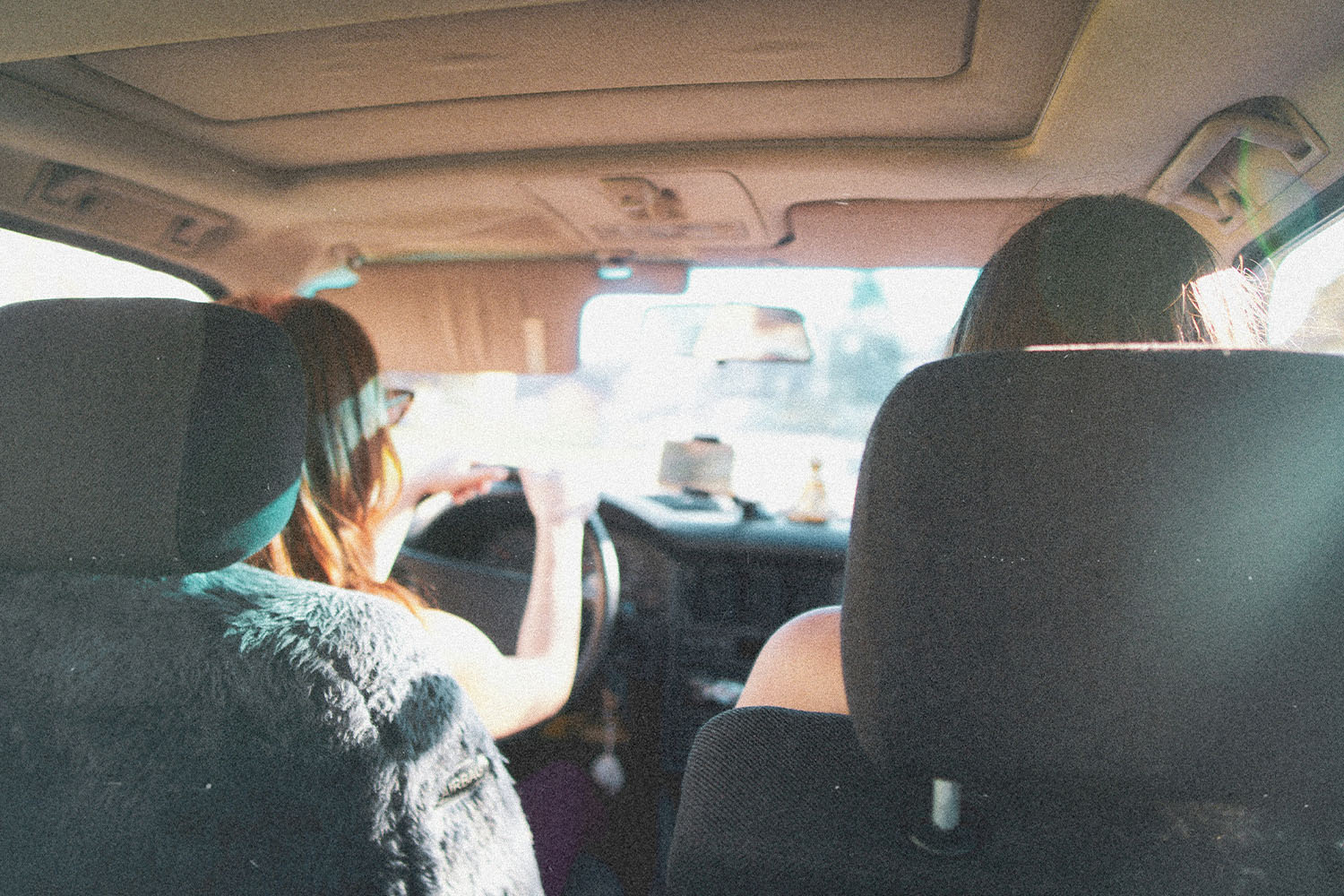 Two people sit in the front seats of a car during daylight, one driving as sunlight streams through the windshield