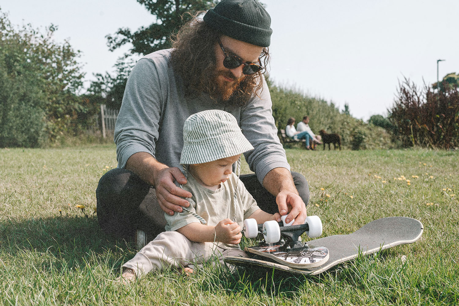 Father helping a baby examine a skateboard while sitting on the grass