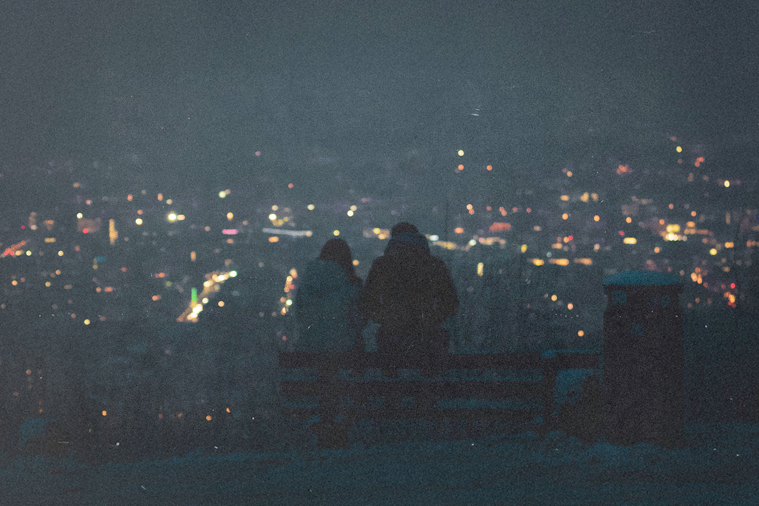 Two people sit on a bench at night overlooking a city filled with glowing lights in the distance