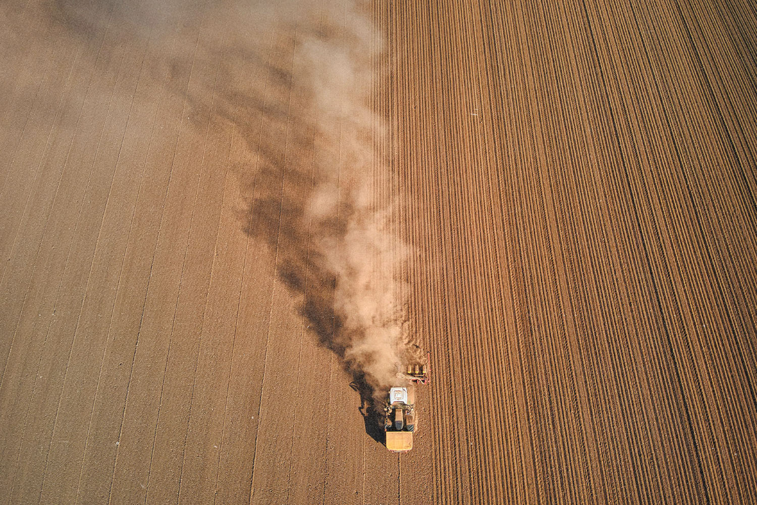 A tractor moves across a large plowed field, leaving a long trail of dust behind it