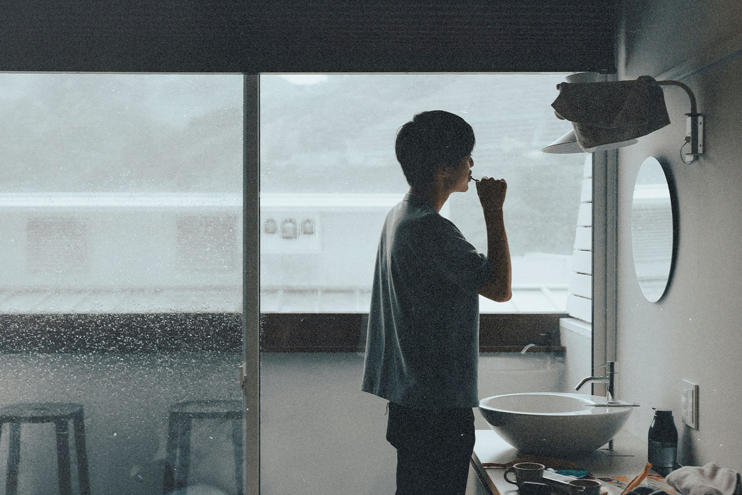 Man brushing their teeth in a small bathroom with a large window overlooking a rainy landscape