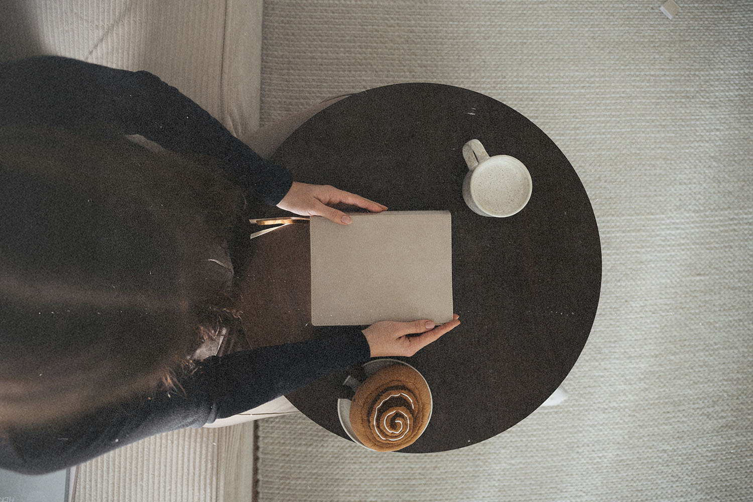 A person sits at a small round table with a closed notebook, a mug of coffee, and a pastry in warm natural light