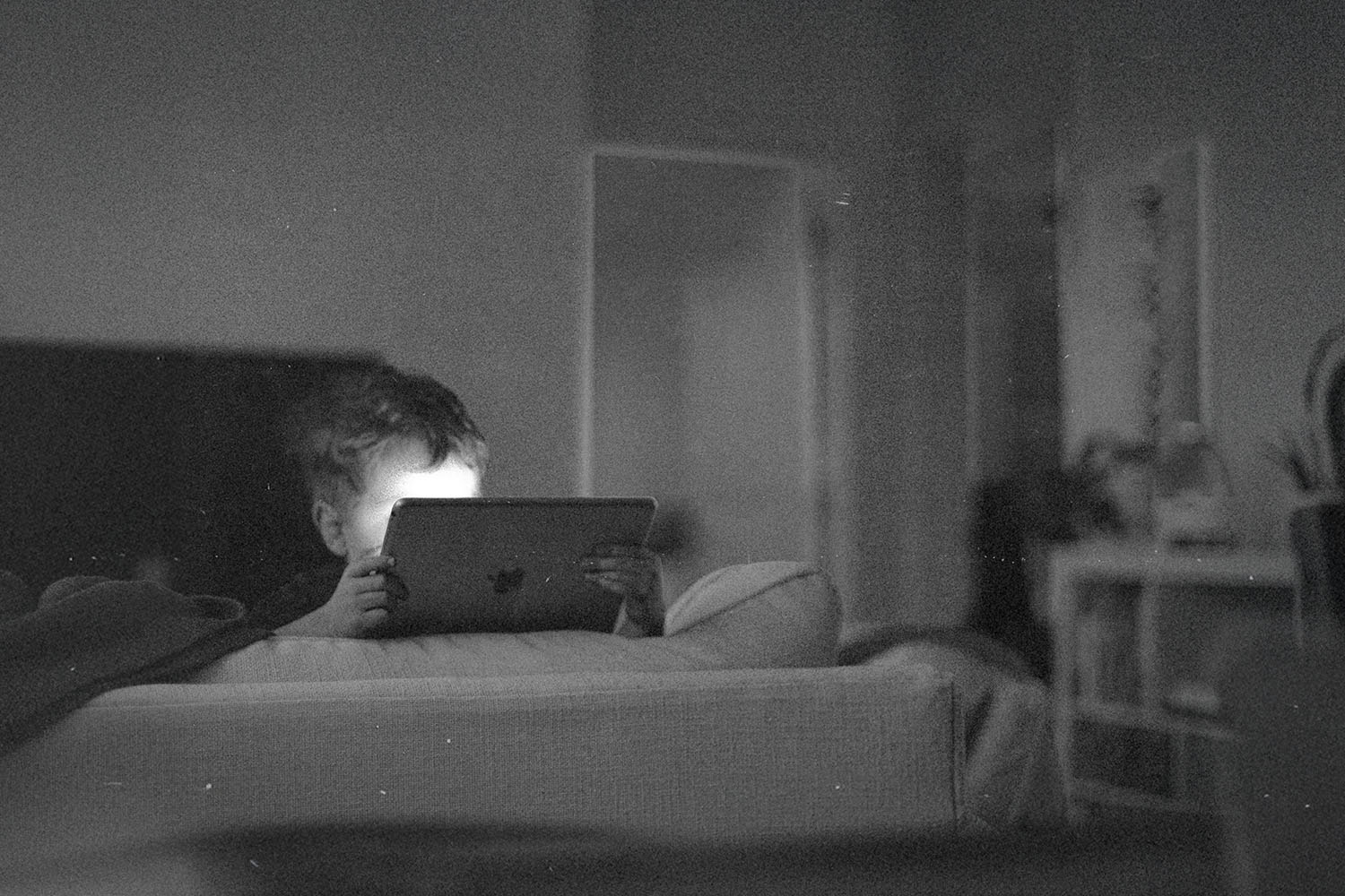 A child sits on a couch in a dim room, holding a tablet with soft light reflecting from the screen