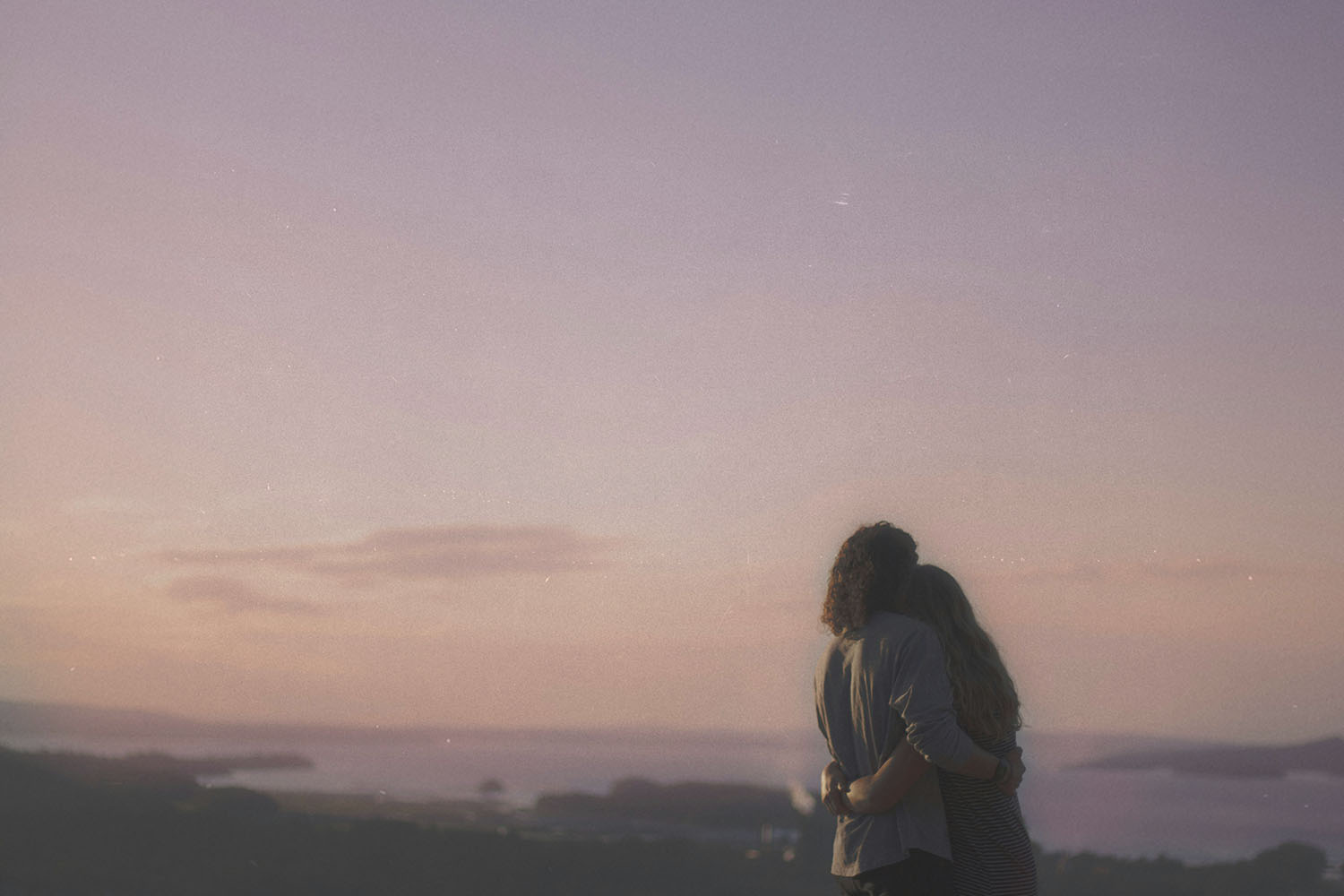 Couple standing arm in arm overlooking a calm lake and distant hills at sunset