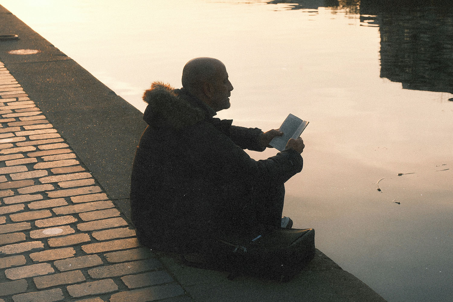 Person sitting on a riverside ledge at sunset, reading a book with calm water reflecting the warm light