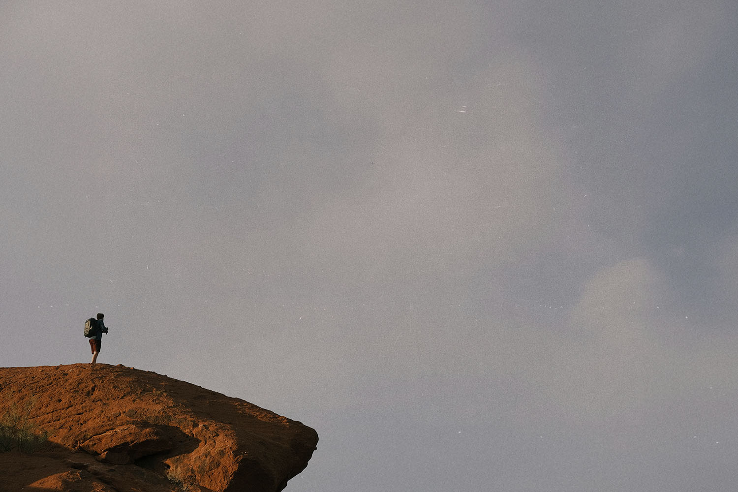 A lone hiker stands on the edge of a rocky cliff under an expansive gray sky