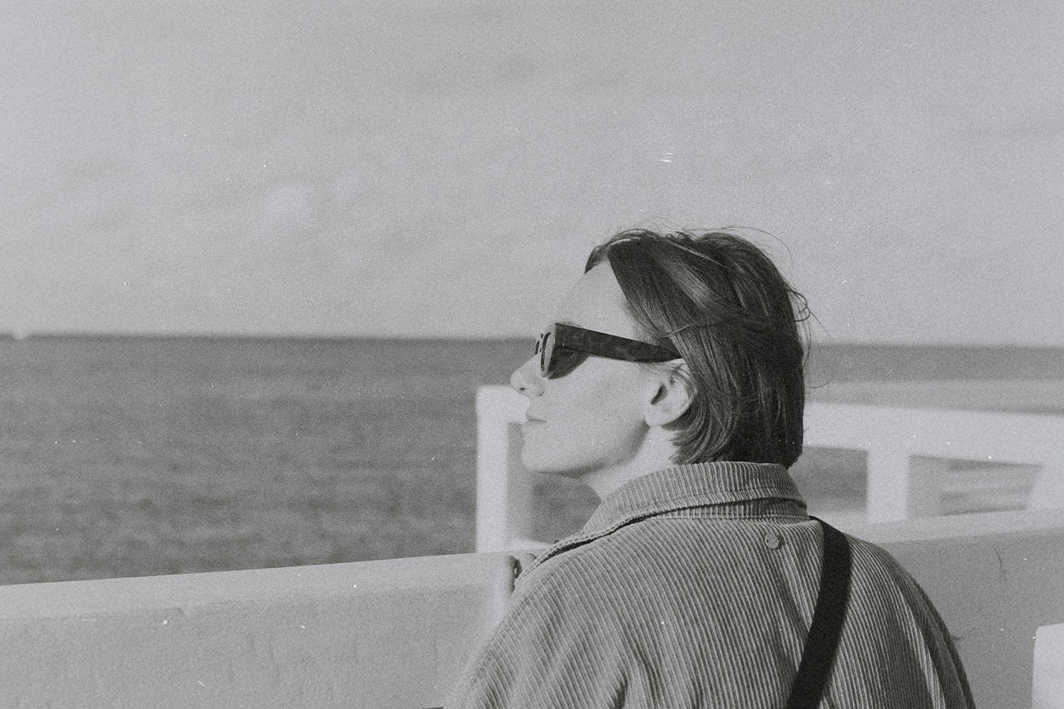 Person standing at a waterfront railing, looking out toward the open sea on a calm, overcast day
