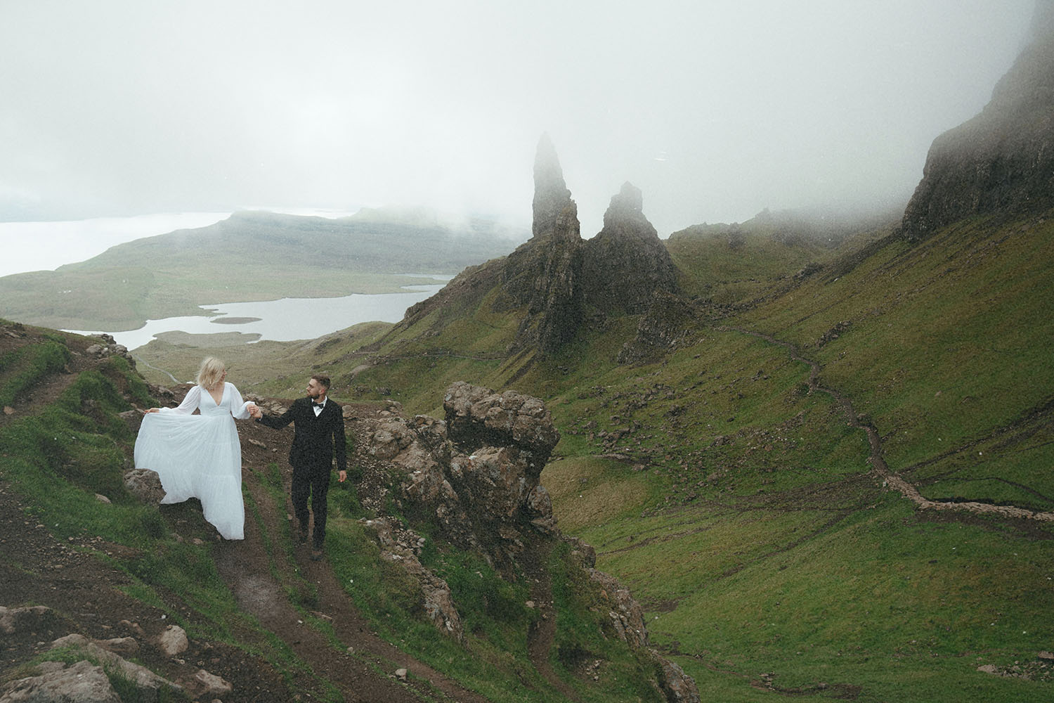 ride and groom holding hands while hiking through mist-covered highlands with steep cliffs and rolling green hills