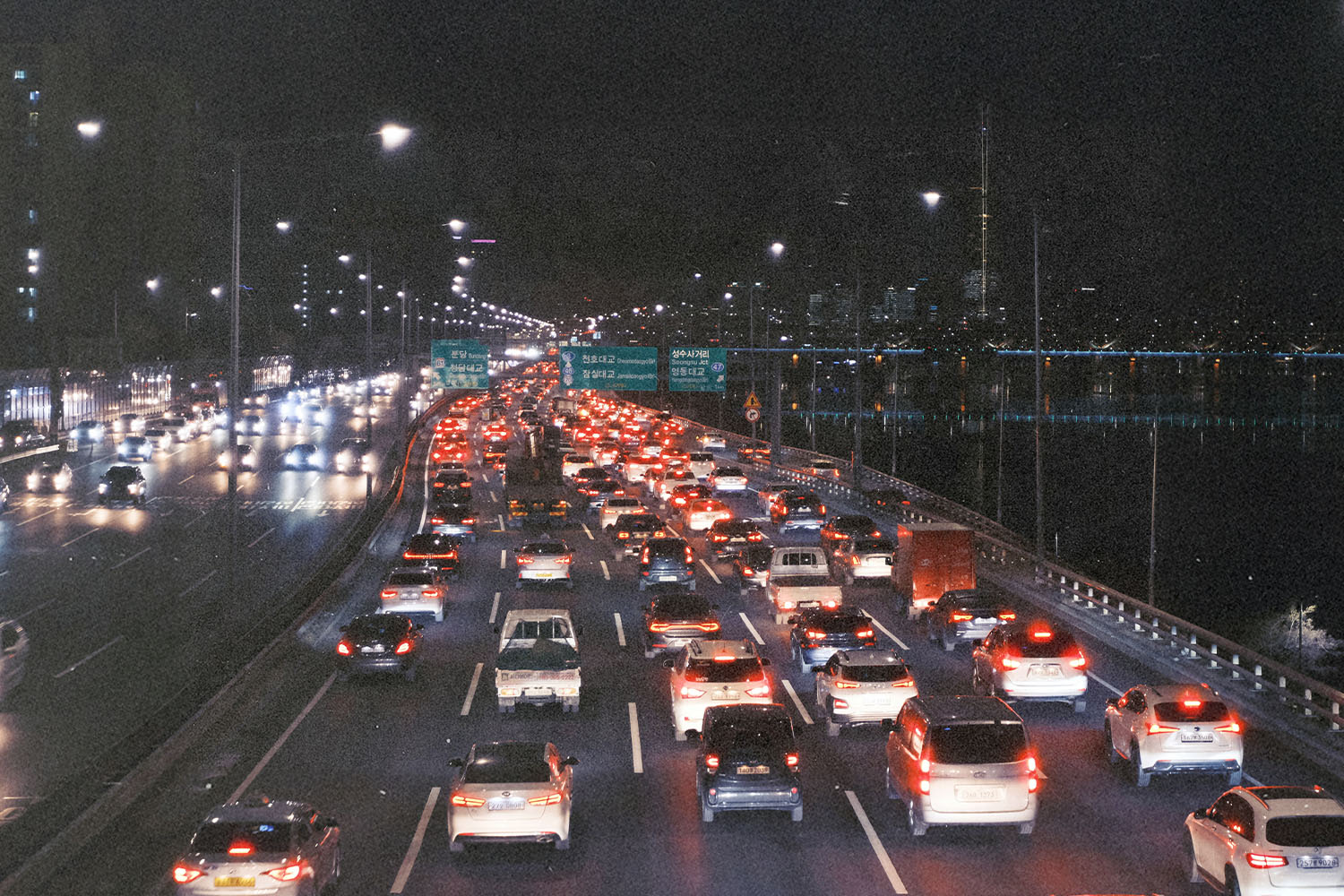Heavy nighttime traffic on a multi-lane highway, with rows of brake lights stretching into the distance beside a riverside cityscape