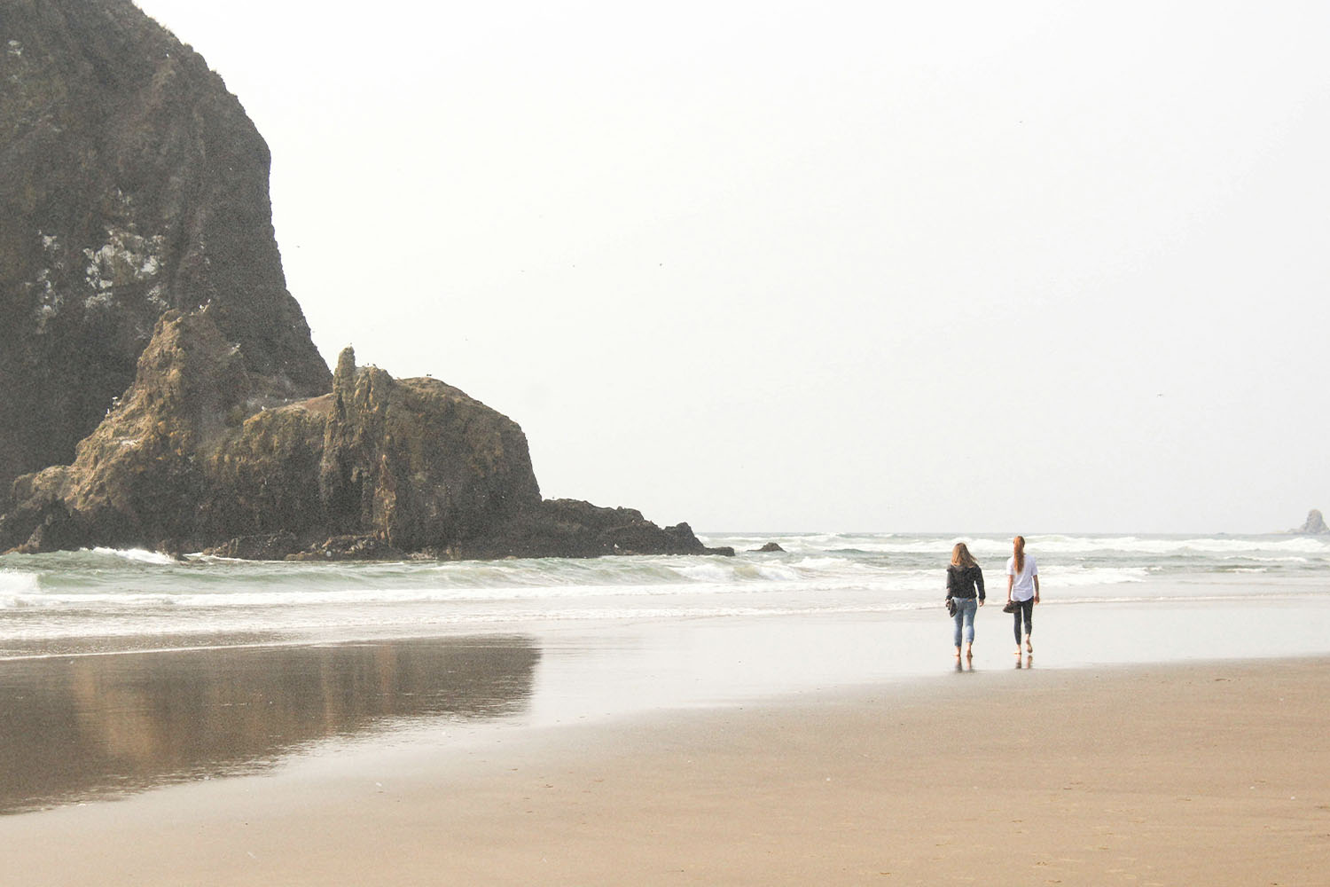Two people stroll along a wide beach with waves rolling in and a large rocky formation rising from the shoreline nearby
