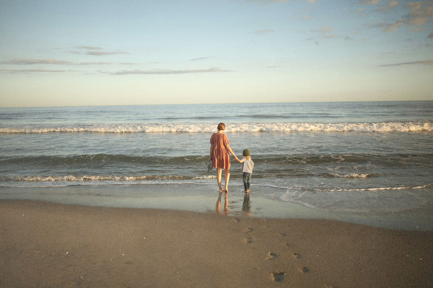 Adult and child standing side by side on the beach, looking out at the calm ocean under a pastel sky