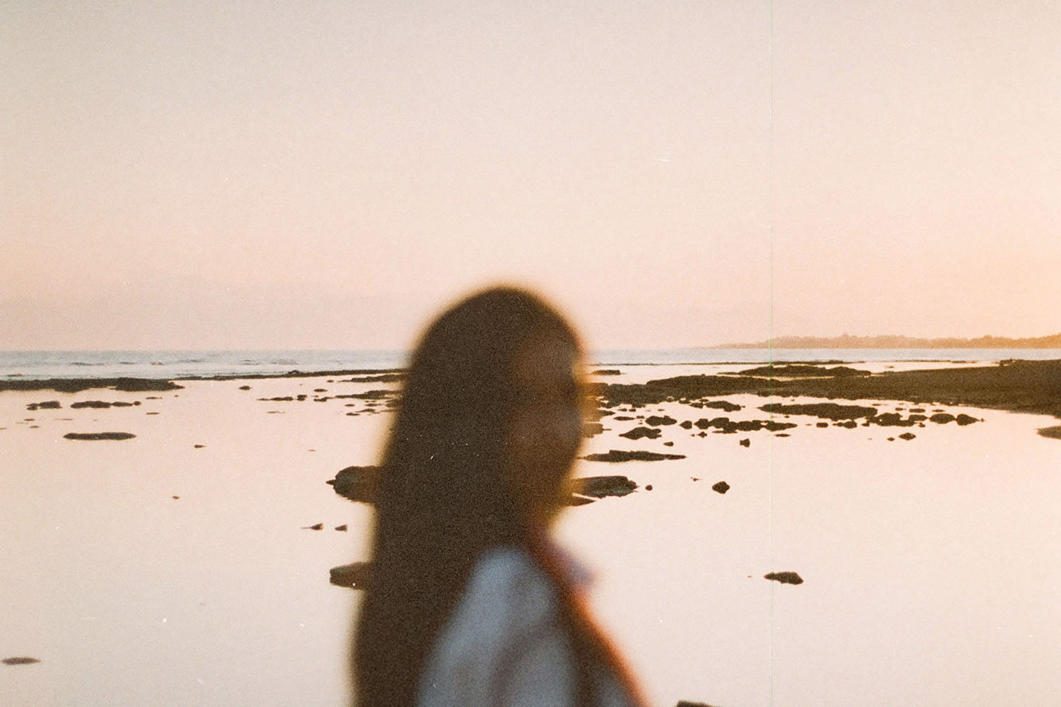 A person stands near the shoreline at sunset, with calm water and scattered rocks stretching into the distance