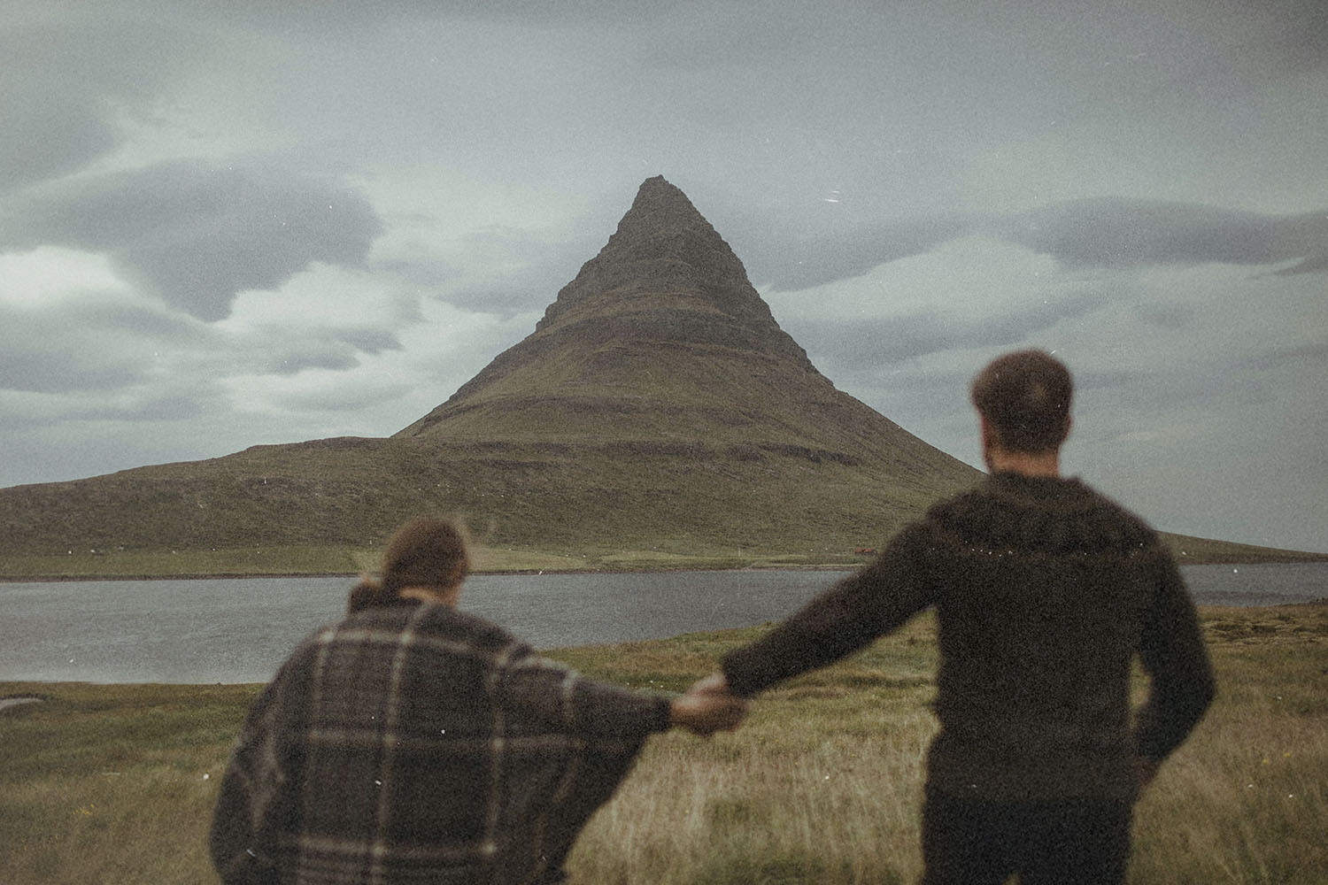 Two people hold hands while facing a tall, cone‑shaped mountain beside a calm body of water